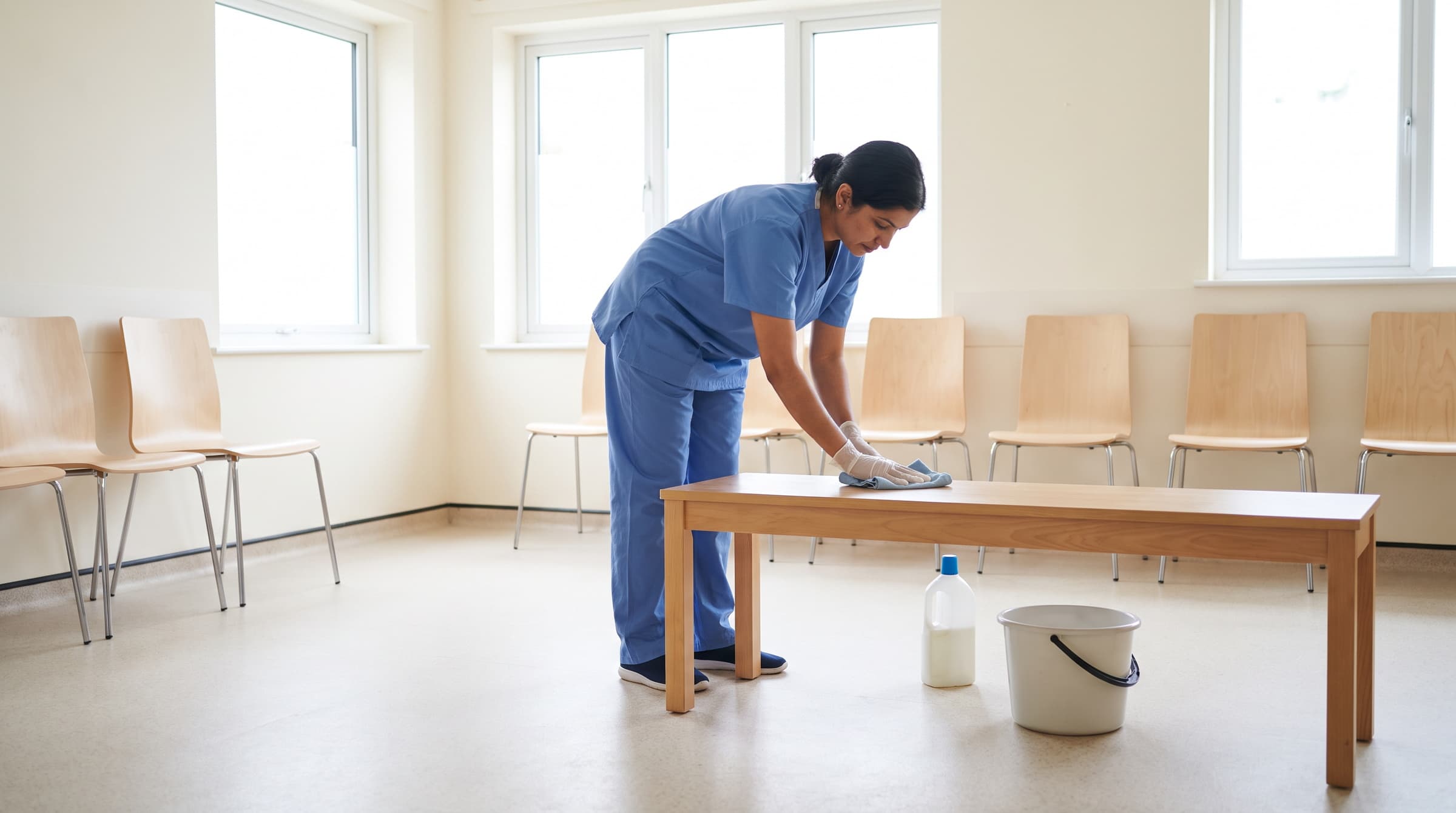 Cleaner in scrubs disinfecting a bright GP surgery waiting room during medical practice cleaning in Devon